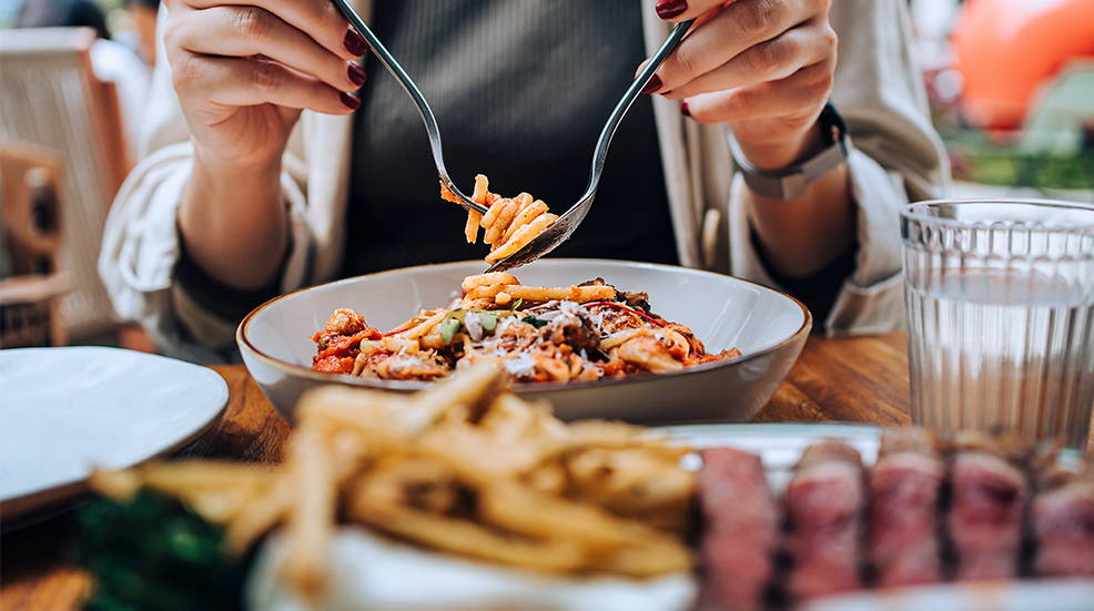 Close-up, mid-section of young woman sitting at dining table holding cutleries, twirling freshly served spaghetti bolognese